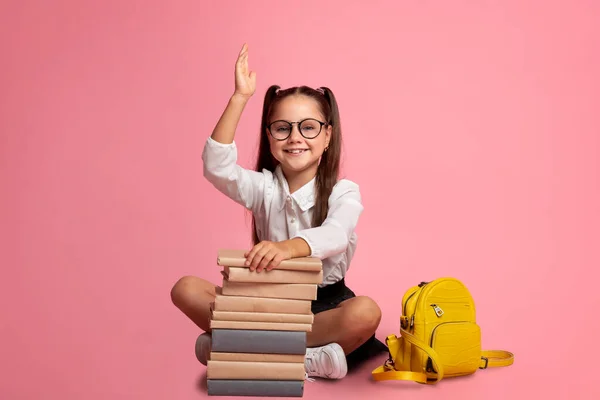 Smart elev. Leende flicka i glasögon med hög med böcker och ryggsäck vet svaret och höjer handen upp — Stockfoto