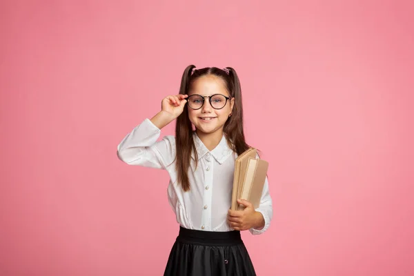 Porträtt av söt liten flicka på rosa bakgrund. Skolflicka tittar på kameran, håller i boken och rätar ut glasögon — Stockfoto