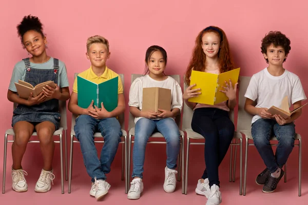 Retrato de comprimento total de crianças em idade escolar com materiais educacionais sentados em cadeiras no fundo rosa — Fotografia de Stock