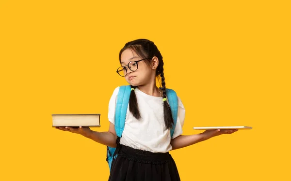 Запутавшаяся японская школьница сравнивает планшет и книгу на жёлтом фоне — стоковое фото