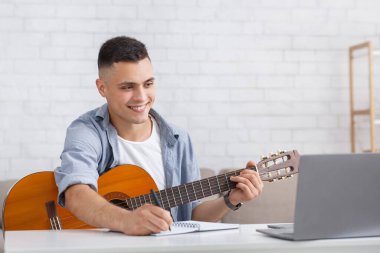 Gülümseyen genç adam çevrimiçi ders izliyor, gitar çalıyor ve notlar alıyor.