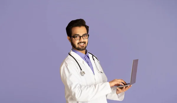 Concepto de medicina cibernética. Feliz médico indio hablando con el paciente en línea en el ordenador portátil, fondo violeta — Foto de Stock