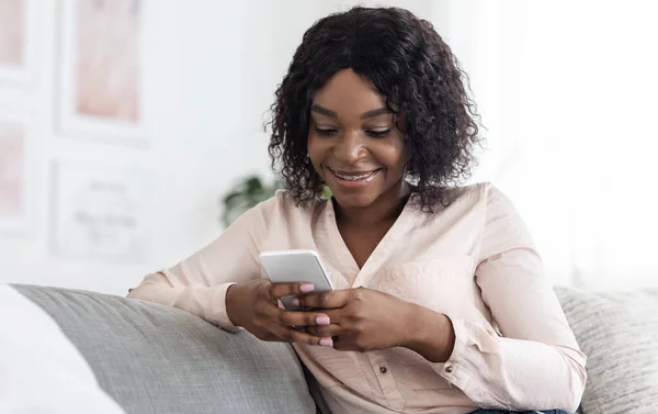 Mulher negra alegre usando telefone celular enquanto relaxa no sofá em casa — Fotografia de Stock