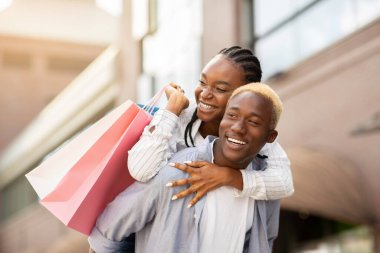 Neşeli Afro-Amerikan milenyumu erkeği renkli çantalı kadını sırtında taşıyor.