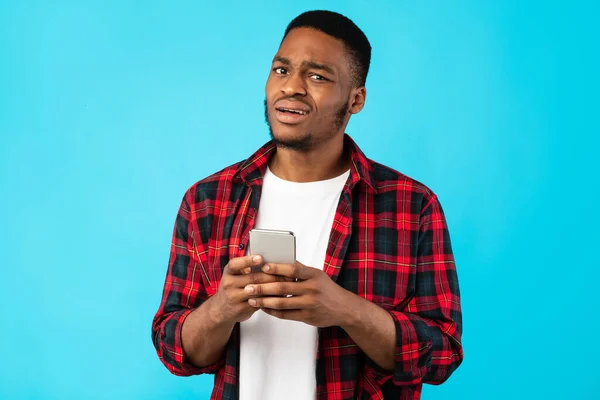 Nejistý černoch drží mobilní telefon přes modré pozadí — Stock fotografie