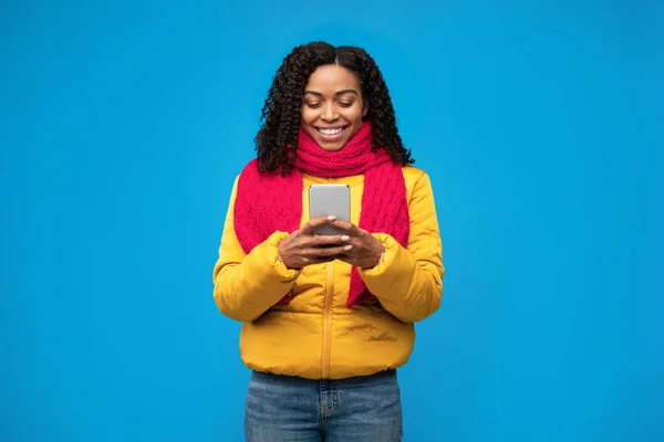 Vrolijk Afrikaans meisje met behulp van telefoon dragen van winterkleding in de studio — Stockfoto