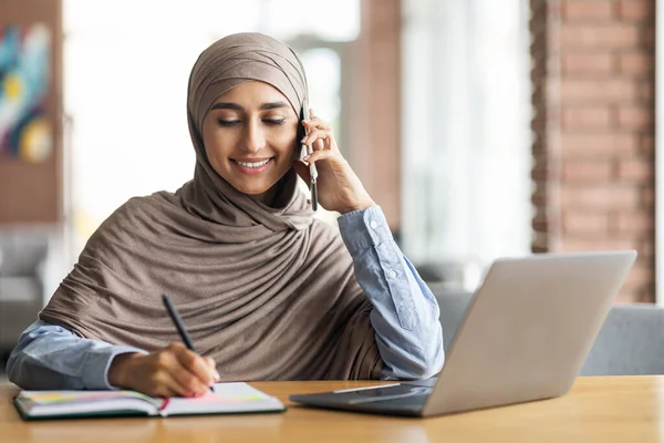 Tesettürlü genç bir kadın telefonla konuşuyor, not alıyor. — Stok fotoğraf