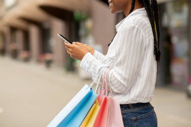 Alışverişe yakın bir yerde buluşacağız. Gülümseyen Afrikalı Amerikalı kadın blogcu. Akıllı telefonuyla yazı yazıyor ve elinde çantalar var.