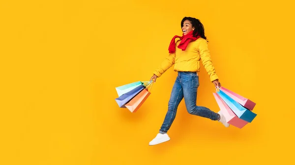 Mujer africana sosteniendo bolsas de compras corriendo en medio del aire, fondo amarillo — Foto de Stock