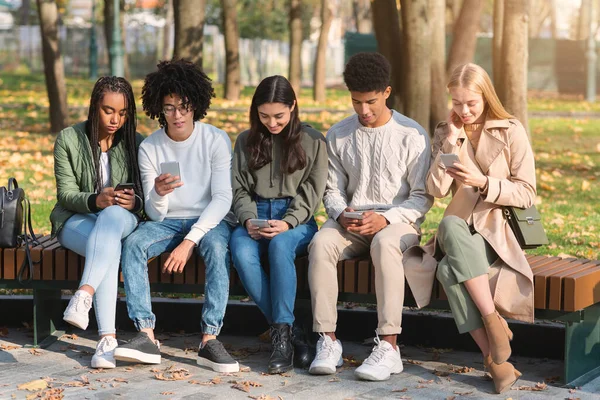 Adolescentes sentados en el banco en el parque y usando teléfonos inteligentes — Foto de Stock