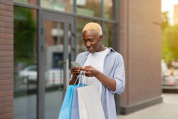 Smiling african american guy with packages with purchases in hands, typing on smartphone