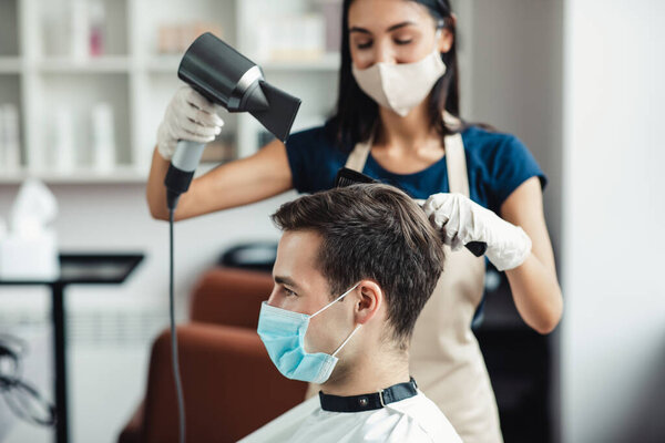 Master in protective mask drying hair of client in mask at salon, close up