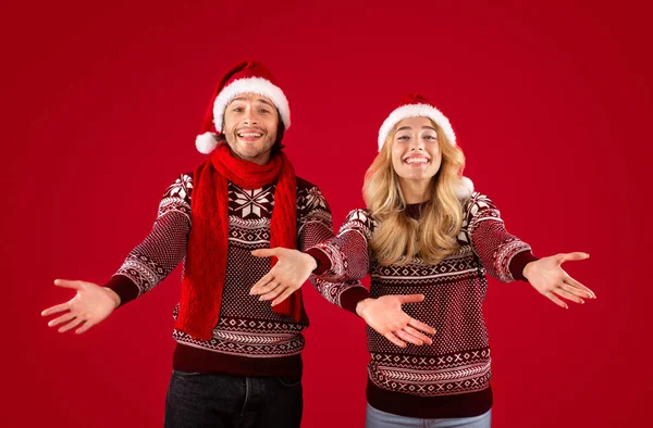 Casal jovem e hospitaleiro em roupas de Natal abrindo os braços em gesto de boas-vindas sobre fundo vermelho — Fotografia de Stock