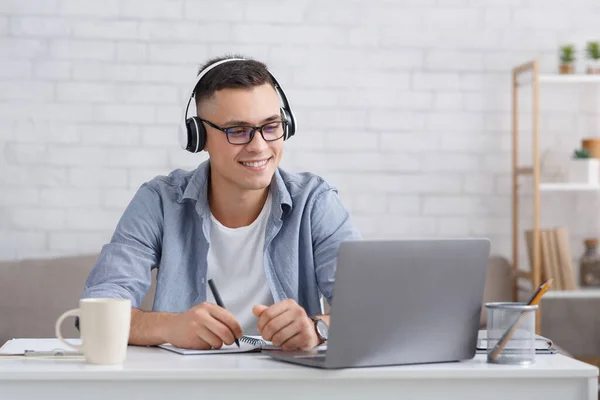 Nuevos conocimientos en línea en casa. Hombre feliz en gafas y auriculares toma notas y mira a la computadora portátil — Foto de Stock
