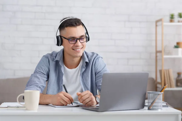 Ny kunnskap på nettet hjemme. Lykkelig mann med briller og hodetelefoner gjør notater og ser på laptop – stockfoto