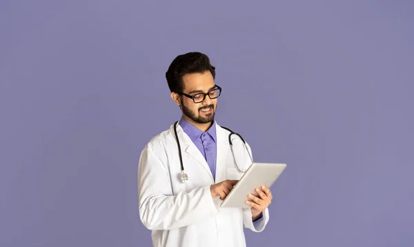 Guapo médico indio con tableta que ofrece consulta en línea al paciente sobre fondo lila — Foto de Stock
