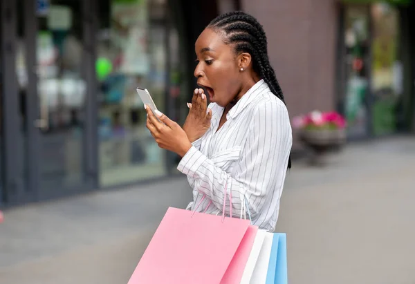 Announcement of big discounts in mobile application. Shocked african american girl with bags received message on smartphone