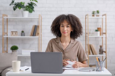 Alternatif işler evde. Not defteri olan tatlı Afro-Amerikan kadın dizüstü bilgisayara bakıyor.