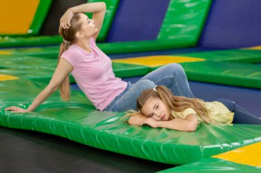 Çocuk eğlence merkezinde yorgun bir kız ve annesi trambolinde dinleniyor.