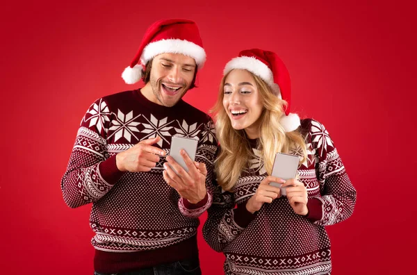 Šťastný tisíciletý pár v Santa klobouky nakupování pro vánoční dárky on-line na červeném pozadí — Stock fotografie