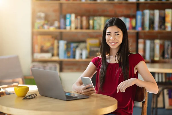 Chica sonriente sentada con el ordenador portátil y el teléfono inteligente en el café, disfrutando de trabajar a distancia —  Fotos de Stock