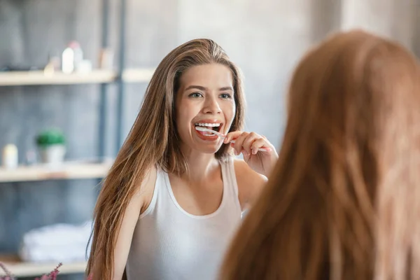 Kebersihan mulut. Wanita yang menarik membersihkan giginya di dekat cermin di kamar mandi — Stok Foto