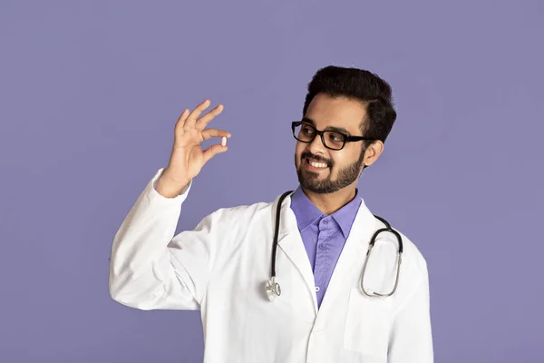 Médico indio sonriente con bata de laboratorio sosteniendo píldora sobre fondo violeta — Foto de Stock