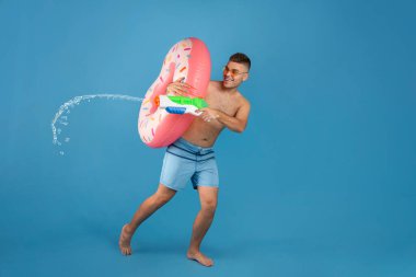 Yaz tatili. Mayo giyen genç adam şişme yüzük tutuyor ve mavi arka planda su tabancası kullanıyor, boşluğu kopyala