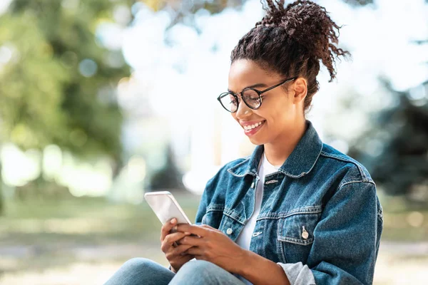 Czarna dziewczyna ze smartfonem Korzystanie z aplikacji mobilnej siedzi w parku — Zdjęcie stockowe