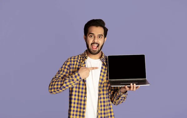 Homem indiano animado apontando para a tela do laptop vazio sobre fundo violeta, mockup para o design do site — Fotografia de Stock