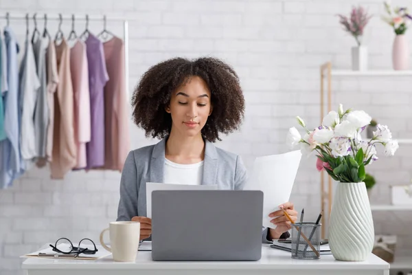 Showroom y oficina en casa. Chica afroamericana ocupada trabajando con documentos cerca de la computadora portátil — Foto de Stock