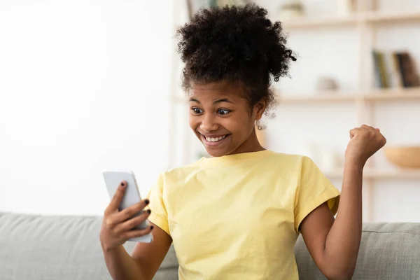 Schwarzes Mädchen gewinnt Spiel auf Smartphone auf Sofa im Haus — Stockfoto