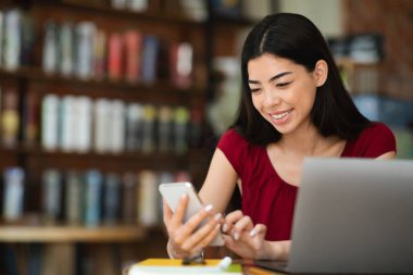Kafeteryadaki dizüstü bilgisayarda çalışırken akıllı telefon kullanan güzel Asyalı kadın.