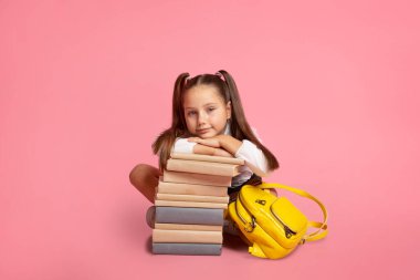 Küçük liseli kız için yeni bilgi. Sırt çantalı çocuk kitap yığınının yanında oturuyor