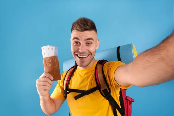 Joven feliz con mochila, pasaporte y pasajes de avión tomando selfie sobre fondo azul — Foto de Stock