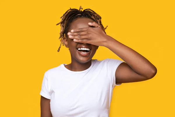 Menina negra feliz cobrindo seus olhos no fundo amarelo — Fotografia de Stock