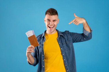 Heyecanlı milenyum adamı mavi arka planda pasaport ve uçak biletlerini gösteriyor.