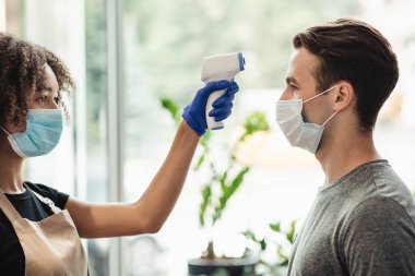 Müşterinin alnının vücut ısısını ölçen güzellik salonu müdürünün yan görüntüsü