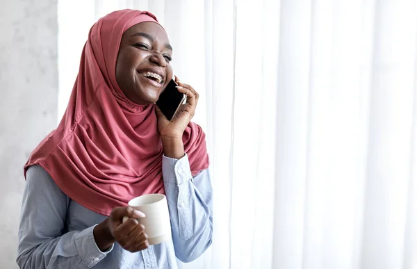 Joyful Black Muslim Woman Drinking Coffee And Talking On Cellphone Near Window — Stock Photo, Image