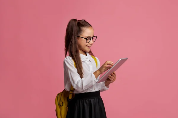 Applicazioni per l'istruzione. Pupilla di scuola elementare in occhiali in uniforme e con zaino, digitando su tablet digitale — Foto Stock