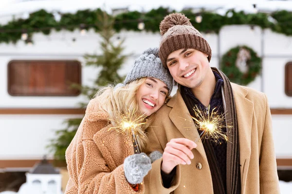 Sonriente pareja romántica en sombreros de punto posando con chispas en el campamento de invierno —  Fotos de Stock