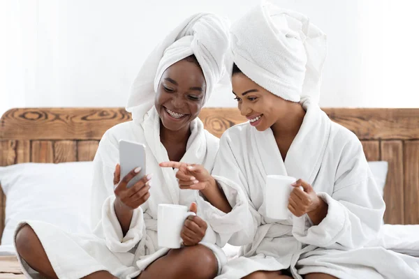 Gelukkig zwarte vrouwen in badjassen het drinken van koffie, met behulp van smartphone — Stockfoto