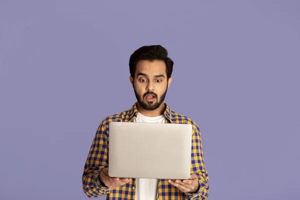 Schockierter Inder starrt mit offenem Mund auf Laptop-Bildschirm vor fliederfarbenem Hintergrund — Stockfoto