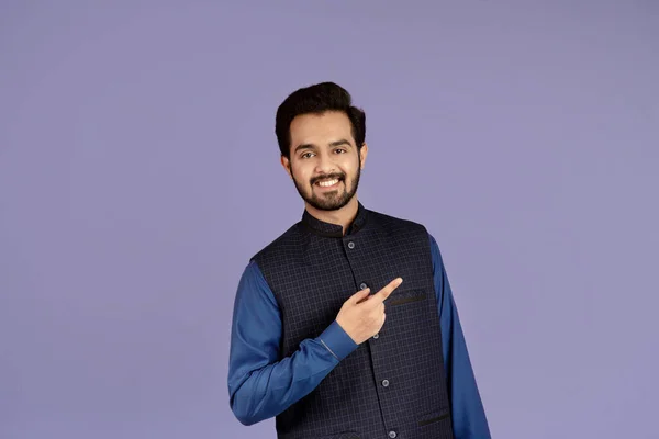 Handsome young Indian man smiling and pointing aside over lilac background