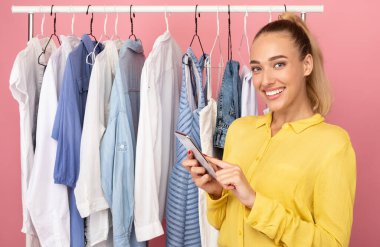 Telefonu elinde tutan mutlu kadın elbise rafının yanında duruyor.