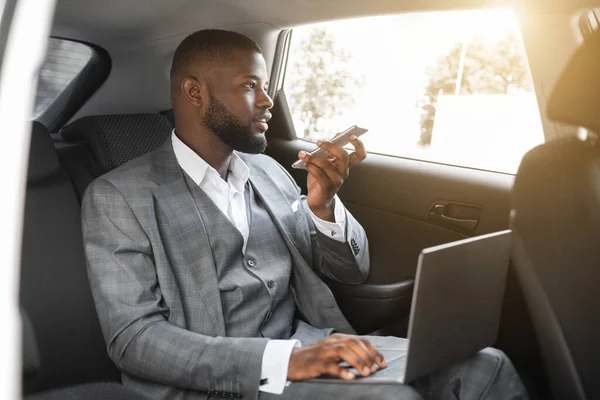 Schwarzer Manager arbeitet am Laptop im Auto und telefoniert — Stockfoto