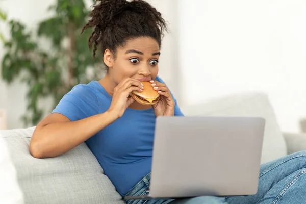 African Girl Eating Burger Using Laptop Sitting On Couch Indoor — Stock Photo, Image