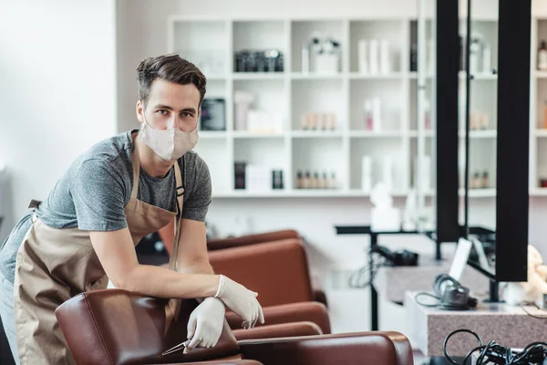 Male hairdresser in protective mask and gloves waiting for clients in empty salon interior