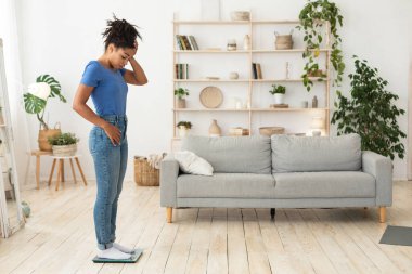 Üzgün Afrikalı kız tartıların üzerinde duruyor, ağırlığını evde koyuyor.