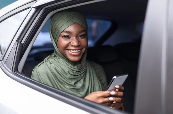 Mujer musulmana negra sonriente sentada en el asiento trasero en el coche, usando un teléfono inteligente — Foto de Stock